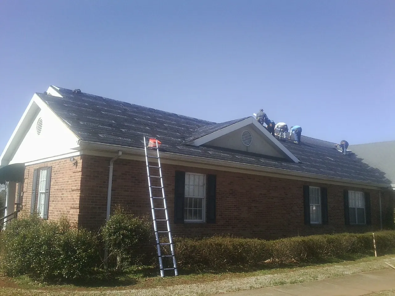 Crew tearing shingles off a brick ranch roof with a ladder set — Emergency Roof Repair work by our crew in Dayville