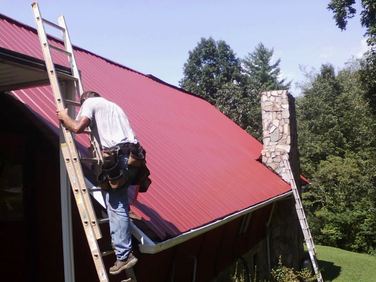 Roofer on a ladder beside a red metal roof and stone chimney — Soffit & Fascia Repair by Roofing Brothers in Dayville
